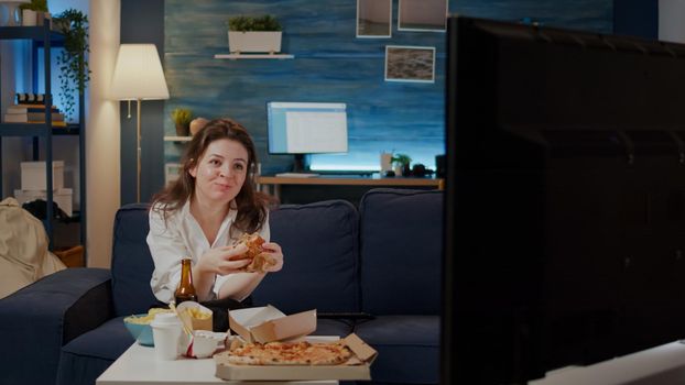 Person eating delicious hamburger and fries in living room while drinking alcoholic beverage and watching television. Woman with takeaway fast food meal sitting on couch and relaxing