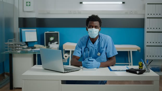 Portrait of young person working as nurse in cabinet at healthcare facility. Medical assistant sitting at white desk looking at camera with face mask, gloves, uniform and stethoscope