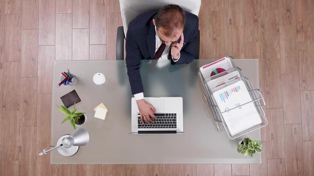 Top view of businessman in suit discussing online profit with manager at phone typing company investments on laptop working in startup business office. Corporate leader analyzing financial graphs