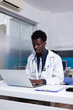 Portrait of african american doctor sitting at desk using laptop computer for examination in medical cabinet at healthcare clinic. Black man with modern device and document files on table