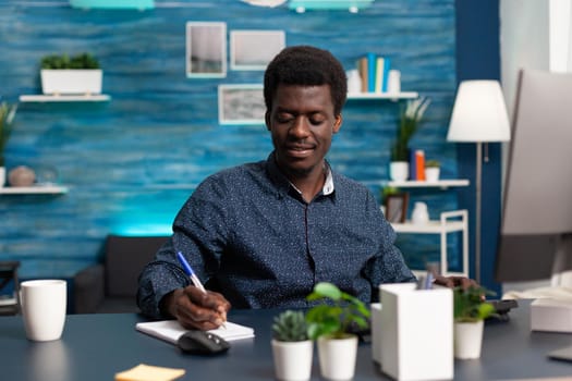 Freelancer taking notes on notebook looking at computer on home office desk. African american man remote working from home and doing online college courses for social media business