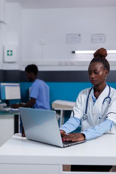 Portrait of african american woman with doctor profession sitting at desk while using laptop and technology in healthcare office. Black medic with modern device and man nurse in background