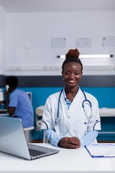 Portrait of african ethnicity doctor looking at camera while sitting at white desk with laptop, documents, and tools. Black medic wearing medical uniform and stethoscope at clinic