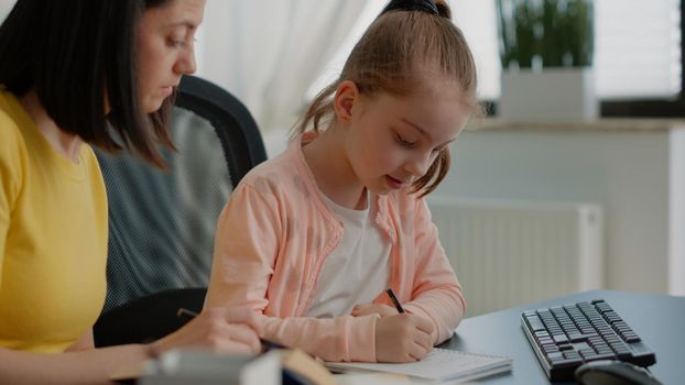 Young child writing homework on notebook and mother assisting for school tasks and learning. Parent helping schoolgirl with exercises on textbook and online remote courses on computer