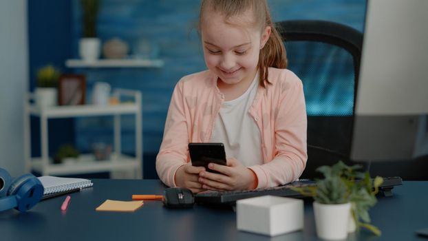 Cheerful child looking at smartphone with touch screen for online classes and lessons. Elementary school girl using mobile phone at desk for remote education and courses with teacher