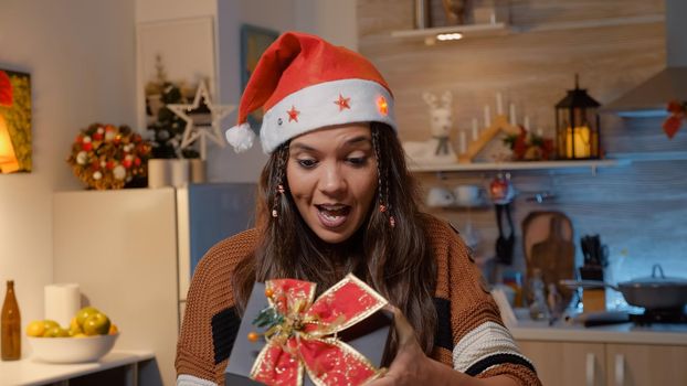 Festive woman with santa hat receiving gift on video call from family in holiday decorated kitchen at home. Young adult being happy while opening present for christmas eve festivity