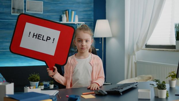 Little child holding speech bubble for help with homework. Schoolgirl sitting at desk with cartoon sign in hand while having school tasks and work for online remote lessons in class.