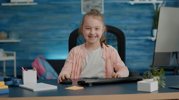 Portrait of schoolgirl at desk using computer and keyboard for online school courses and homework. Little child smiling and looking at camera while having technology for remote education