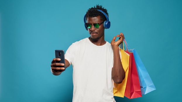 Playful man taking pictures with shopping bags on smartphone while wearing sunglasses and headphones. Modern adult taking selfies with phone after clothing purchase from retail.