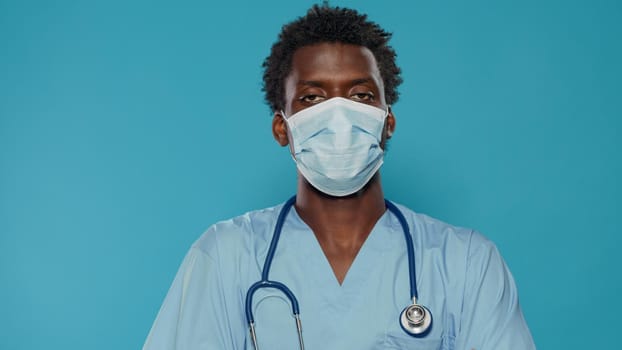 Close up of medical assistant with face mask looking at camera in studio. Portrait of man working as nurse, wearing blue uniform and stethoscope while having protection against covid 19