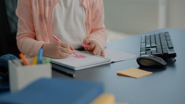 Close up of young child using colorful pencils on notebook at desk. Little girl holding pen to draw doodle on textbook while sitting at home after school homework. Schoolgirl drawing