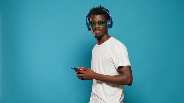 Playful adult dancing to song rhythm on headphones in studio, looking at camera. Cheerful man listening to music using smartphone and headset while wearing sunglasses over isolated background