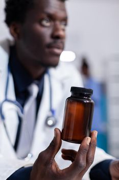 Close up of african american hand with bottle of pills for treatment recommendation. Black doctor holding flask with medicine, medicament and drugs. Medic with vial in medical cabinet