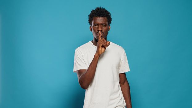 Young man doing silence gesture with finger on mouth in studio. Confident person asking for secrecy and quiet, using forefinger to make censorship expression over isolated background.