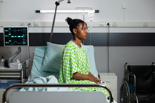 Young african american person sitting on hospital ward bed with fever, sickness, disease. Sick teenager with oximeter on hand, medical equipment and heart rate monitor for recovery