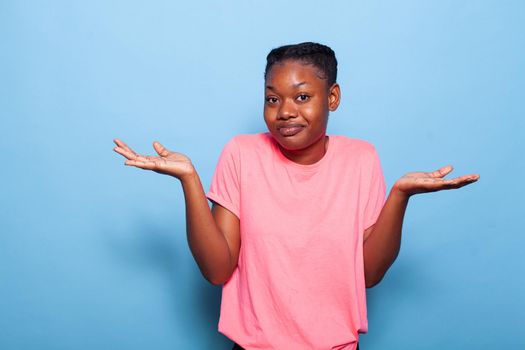 Portrait of confused african american teenager posing in pink t-shirt on a blue background in studio making uncertain facial expression. Young woman shrugging shoulders. I dont know concept