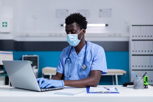 Black adult with nurse profession working at healthcare clinic using laptop and modern technology for medical system. African american man wearing uniform sitting at desk during pandemic