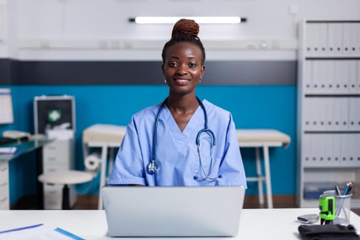 Portrait of african american nurse using laptop at white desk sitting in medical office. Black woman with assistant occupation wearing uniform and working at healthcare clinic