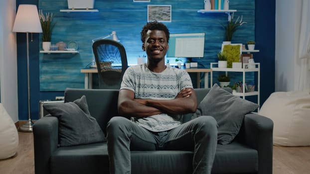 African american adult sitting on couch and looking at camera. Portrait of black person with crossed hands smiling while being at home with technology. Man of african ethnicity