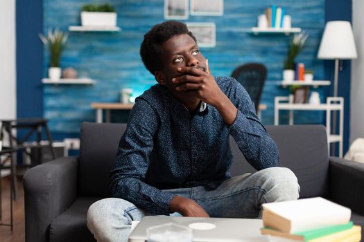 Portrait of thoughtful african american man looking out the window, thinking about workplace business problems. Black adult sitting in living room on couch working from home office