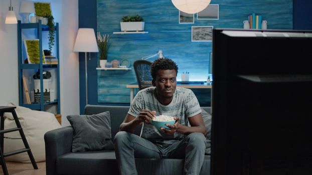 Black adult watching comedy movie on television while eating popcorn from bowl. African american person laughing and looking at TV with snack. Man of african ethnicity having fun