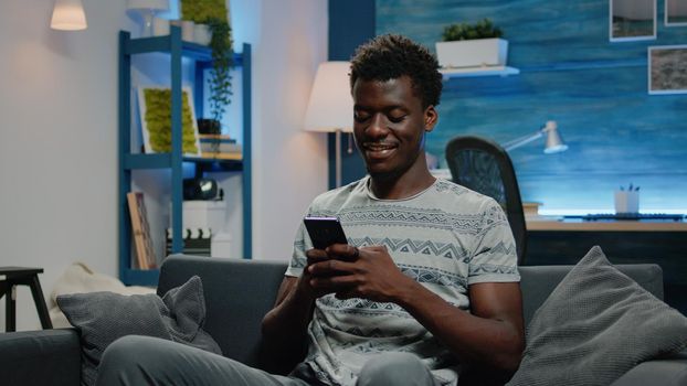 African american man smiling and using smartphone to text messages for online remote work. Black entrepreneur texting on phone while sitting on couch. Person holding touch screen device