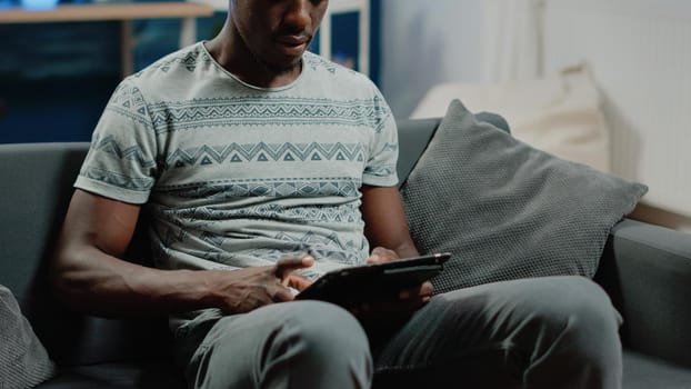 Close up of african american man typing on digital tablet with touch screen after remote business work. Black person using gadget as free time activity on couch. Freelancer using device