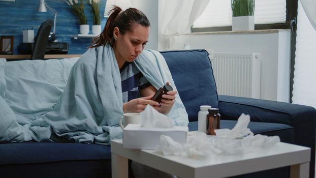 Person reading label of tablet with capsules and bottle of pills against virus infection. Woman with flu searching for treatment to cure illness and blowing runny nose with tissue.