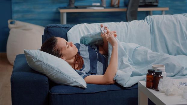 Sick woman resting on sofa and using oximeter for oxygen saturation measurement and to check pulse pressure. Adult with virus disease, medical instruments and healthcare treatment.