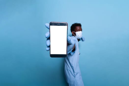 Medical assistant with vertical blank screen on smartphone. Man nurse with covid 19 protection face mask and gloves showing cellphone with white isolated background on empty display