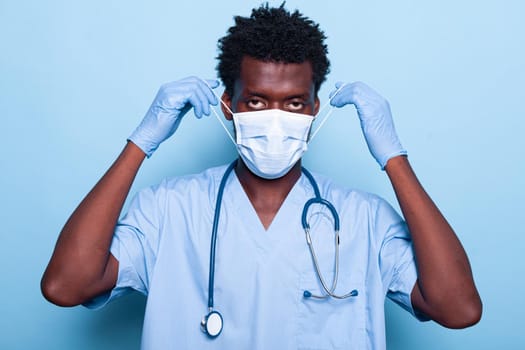 Portrait of medical assistant putting mask on face for protection against coronavirus pandemic. Man working as nurse wearing uniform, stethoscope and gloves while looking at camera