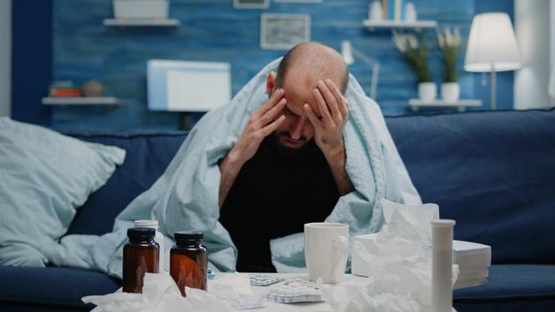 Close up of sick adult with headache rubbing temples in blanket. Man with migraine and flu symptoms trying to cure pain while having medicaments, pills, tea and thermometer on table