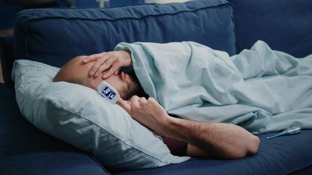 Close up of adult using oximeter for disease diagnosis. Medical device with decreased oxygen saturation on finger of sick person laying on couch. Man checking lower pulse pressure