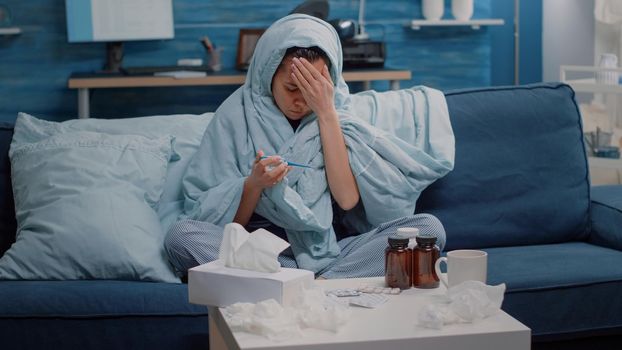 Sick woman covering with blanket and looking at thermometer to check temperature. Adult with cold and flu using medical device, measuring fever to cure illness with medication.