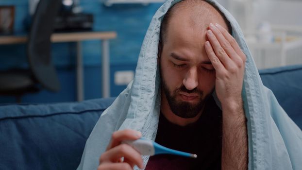 Close up of unwell adult looking at thermometer and measuring temperature while wrapped in blanket. Sick man with illness holding medical instrument to check fever and symptoms.