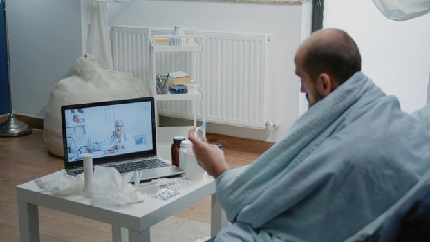 Sick man talking to doctor on video call for telemedicine, holding thermometer with temperature measurement. Patient with fever asking medic on online conference about medical advice