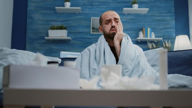 Close up of man with headache holding head and feeling sick. Person with migraine rubbing temples sitting wrapped in blanket while having virus symptoms and healthcare problems.