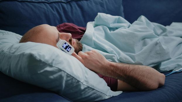 Close up of ill man sleeping on sofa with oximeter on finger. Adult using medical device with lower oxygen saturation measurement. Sick person with flu checking pulse pressure at home