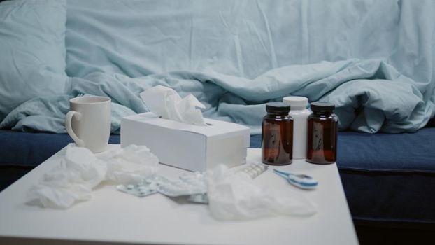 Nobody in living room with medical treatment against flu and seasonal cold. Close up of table with medicaments, medicine, vitamins, bottles of pills, thermometer and cup of tea.