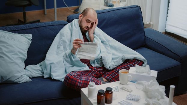 Man with sickness reading medicament information leaflet to heal cold and flu. Person with virus symptoms holding medication paper against disease in blanket while having headache.