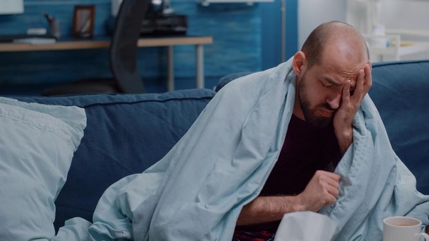 Close up of ill man with headache blowing nose using tissues while having cup of tea and bottles of pills on table. Sick adult with migraine and flu symptoms trying to cure disease