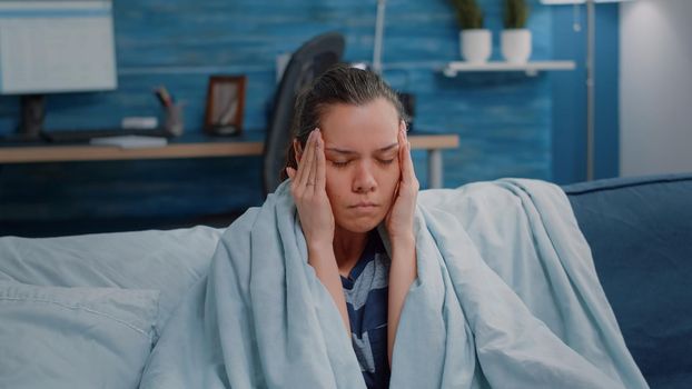 Close up of ill adult rubbing temples against headache. Sick woman with migraine sitting wrapped in blanket, having coronavirus symptoms. Person with virus disease holding hands on head