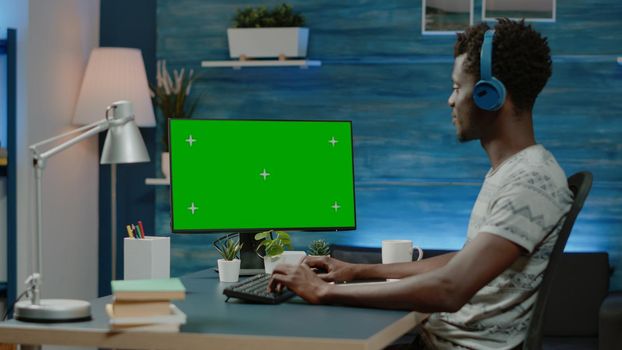 Man looking at horizontal green screen on computer for remote work. Person with headphones working from home, using chroma key on device with mockup template and isolated background.