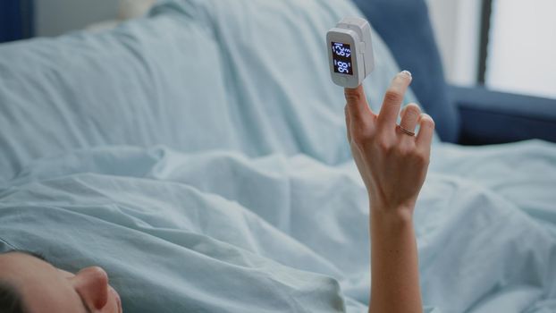 Close up of unwell woman holding oximeter on finger for oxygen saturation measurement and checking pulse pressure while laying on sofa. Person with sickness looking at medical gadget