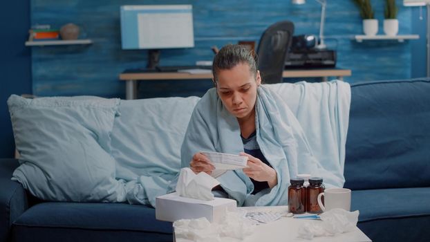 Adult with cold reading medicine leaflet with information to heal seasonal flu symptoms. Sick woman in blanket looking at medicament paper against disease. Person with healthcare problems