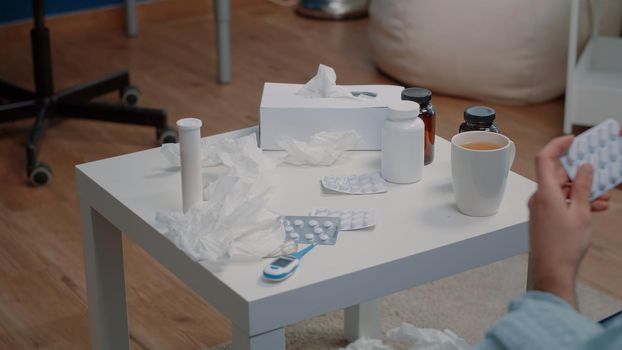 Close up of hand holding tablets of capsules for treatment. Person with medicaments, bottles of pills, medication, thermometer, cup of tea and tissues on table. Adult with prescription medicine