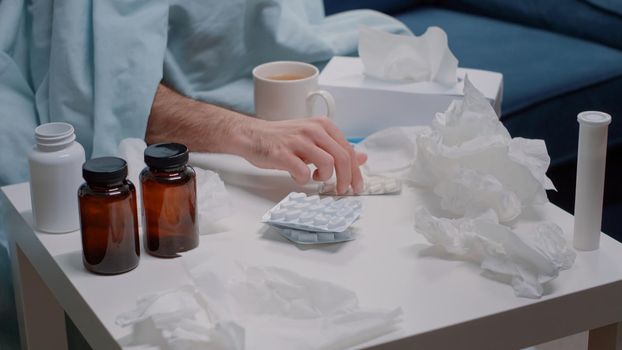Close up of hand searching for capsule tablets against virus symptoms. Adult with bottles of pills, tea, thermometer and tissues on table trying to cure cold and flu. Person with sickness