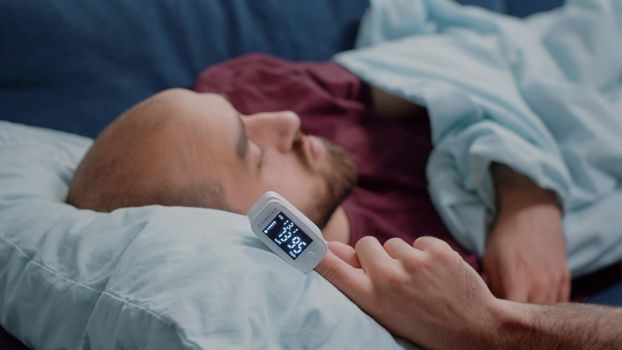 Close up of oximeter on hand of ill person resting while measuring oxygen saturation and pulse pressure to check virus symptoms. Sick adult using device for healthcare treatment