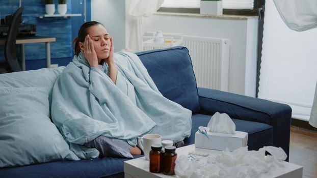 Sick woman rubbing temples and trying to cure headache while sitting wrapped in blanket. Adult with flu symptoms having migraine and virus infection. Person feeling unhappy and ill.