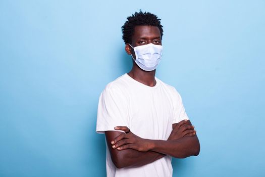 Portrait of person with face mask standing with arms crossed while looking at camera. Casual man with healthcare protection against coronavirus pandemic. Young adult in studio